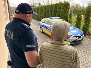 Widziani od tyłu przed wyjściem z domu policjant i starsza kobieta, w tle radiowóz