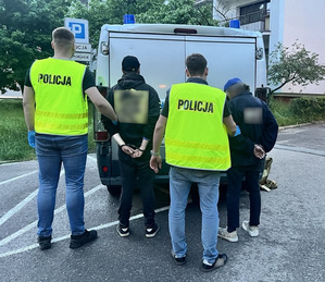 Dwóch umundurowanych policjantów i dwóch mężczyzn ze skutymi kajdankami rękami przed radiowozem typu bus