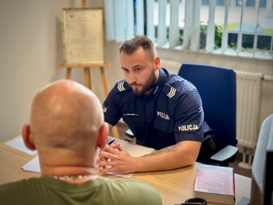 W pomieszczeniu policjant siedzący przy biurku rozmawia z osobą