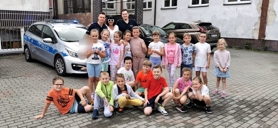 Grupa dzieci oraz wychowawczyni i dwóch policjantów pozujący do zdjęcia na tle radiowozu i budynku