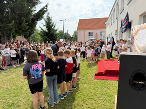 Festyn przed budynkiem szkoły na tle zieleni policjant i grupa dzieci