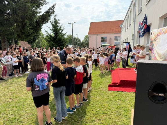 Festyn przed budynkiem szkoły na tle zieleni policjant i grupa dzieci