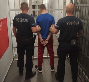 Widziani od tyłu dwaj policjanci przytrzymujący skutego kajdankami mężczyznę w korytarzu aresztu