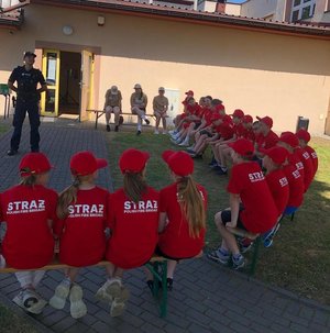 Grupa młodzieży w czerwonych T-shirtach Straży pożarnej siedząca na ławkach na zewnętrz budynku ustawionych wokół policjanta prowadzącego pogadankę