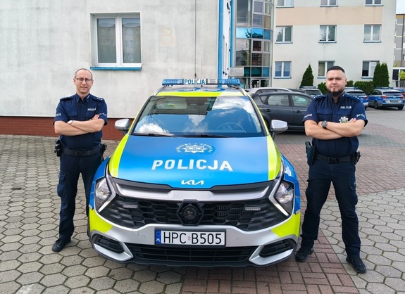 Dwóch umundurowanych policjantów stoi przy radiowozie