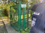 Brama wejściowa na teren ogródków działkowych na pierwszym planie z prawej strony rękaw bluzy policyjnej