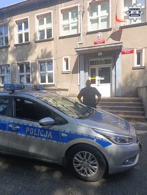 Zdjęcie jednopiętrowego budynku szkoły na pierwszym planie radiowóz, dalej policjant odwrócony tyłem