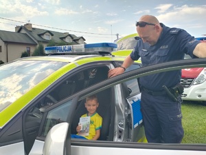 Uśmiechnięty chłopiec siedzący za kierownicą radiowozu, w otwartych drzwiach policjant