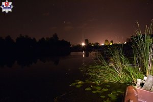 Zbiornik wodny nocą, na pierwszym planie szuwary, w tle światła