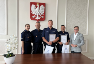 Na tle ściany i godła stojąca czwórka policjantów oraz mężczyzna w marynarce