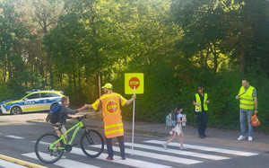 Dziecko przejeżdżające przez jezdnię, policjantka oraz dwaj mężczyźni ubrani w kamizelki odblaskowe