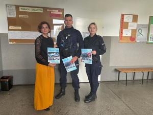 Na tle korytarza szkolnego dwójka policjantów oraz kobieta z plakatami w ręku