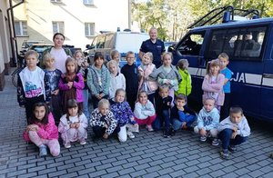 Grupa dzieci z wychowawczynią oraz dwaj policjanci na dziedzińcu komisariatu, w tle radiowozy