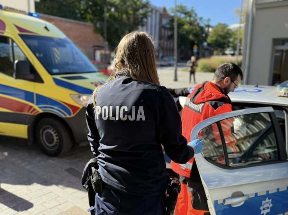 Widziana od tyłu policjantka w głębi radiowóz, karetka i zespół ratownictwa medycznego