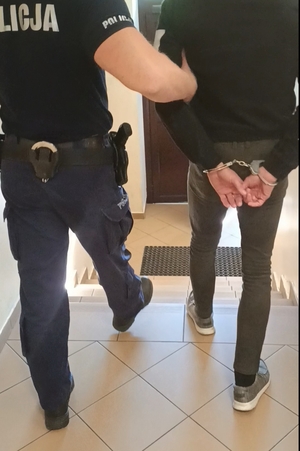 Zbliżenie widzianego od tyłu policjanta prowadzącego korytarzem mężczyznę ze skutymi z tyłu rękami