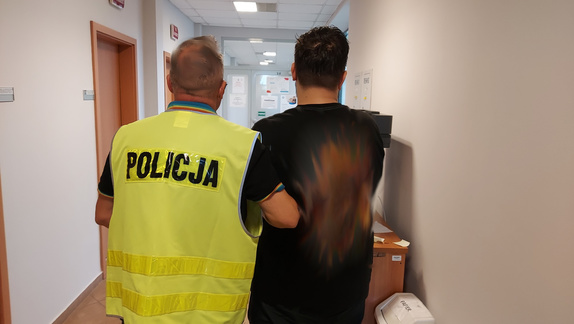 W korytarzu widziani od tyłu mężczyzna w czarnej koszuli przytrzymywany przez policjanta w żółtej kamizelce odblaskowej.