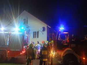 W scenerii nocnej budynek jednorodzinny, wozy strażackie, strażacy i ludzie po cywilnemu