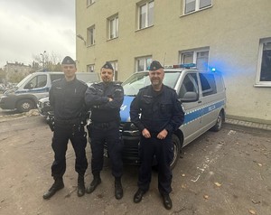 Trzej policjanci na tle radiowozu typu bus i budynku