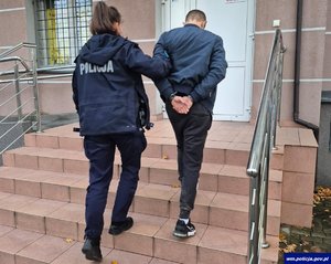 Widziani z tyłu: policjantka wprowadzająca po schodach do komisariatu mężczyznę ze skutymi kajdankami rękami