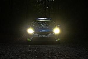 W scenerii nocnej leśnej polany radiowóz z włączonym  oświetleniem