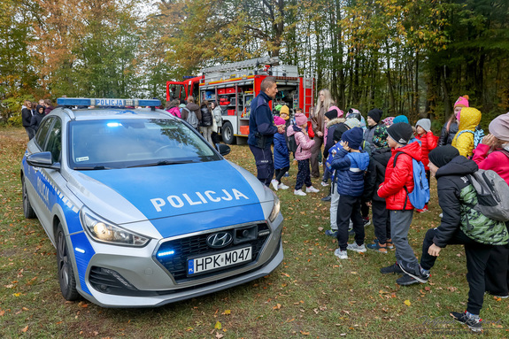 Na leśnej polanie wóz straży pożarnej, radiowóz grupa dzieci oraz policjant i strażak