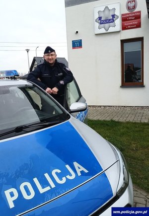 Policjant w drzwiach radiowozu na tle budynku posterunku Policji