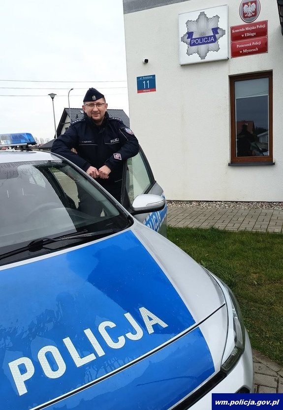 Policjant w drzwiach radiowozu na tle budynku posterunku Policji