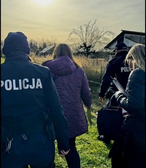 Widziani od tyłu dwaj policjanci oraz dwie kobiety na terenie ogródków działkowych