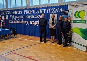 Trójka policjantów oraz kobieta w sali gimnastycznej w tle widoczny  napis &quot;Gminna debata p[profilaktyczna...&quot;