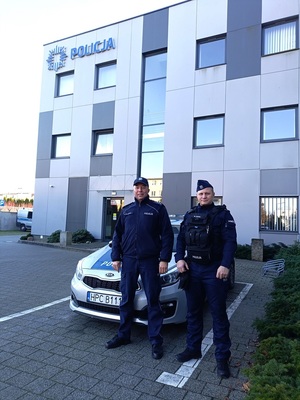 Dwaj policjanci na tle radiowozu i komisariatu