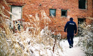 W scenerii zimowej widziany od tyłu policjant idący w kierunku pustostanu z czerwonej cegły