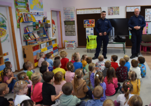 Dwaj policjanci oraz grupa przedszkolaków siedzących na podłodze sali przedszkolnej