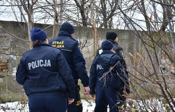 Dwójka policjantów i dwójka strażników widziana od tyłu w scenerii zimowej na tle betonowego ogrodzenia