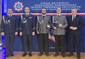 Pozujący do zdjęcia czterej policjanci, z których dwóch w ręku trzyma odznaczenia oraz mężczyzna w garniturze
