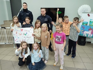 Dwaj policjanci pozujący do zdjęcia z grupką dzieci trzymających narysowany odręcznie plakat z napisem kochaj, szanuj, nie krzywdź, nie bij