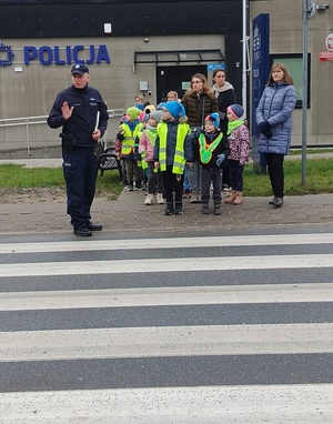 Na tle budynku policji policjant przeprowadzający dzieci przez zebrę