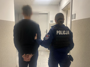 Widziani z tyłu policjantka przytrzymująca w korytarzu mężczyznę ze skutymi kajdankami rękami