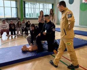 W sali gimnastycznej policjant demonstrujący młodzieży udzielanie pierwszej pomocy