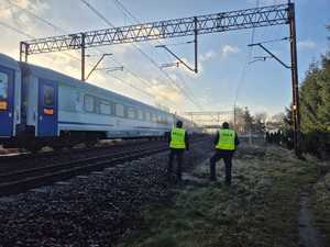 Widziani z tyłu dwaj policjanci w kamizelkach odblaskowych stojący na nasypie kolejowym obok przejeżdżającego pociągu