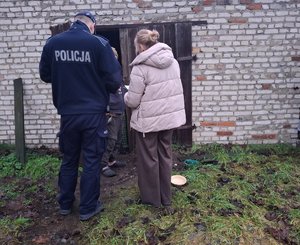 Widziani z tyłu policjant oraz kobieta w kurtce, za nimi mężczyzna w zniszczonej odzieży na tle budynku gospodarczego
