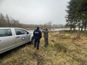 Policjant oraz mężczyzna w zielonej bluzie obok samochodu terenowego na brzegu zbiornika wodnego