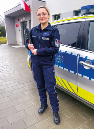 Uśmiechnięta policjantka obok radiowozu na tle posterunku