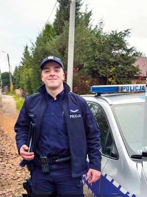 Uśmiechnięty policjant obok radiowozu na tle osiedla domków jednorodzinnych