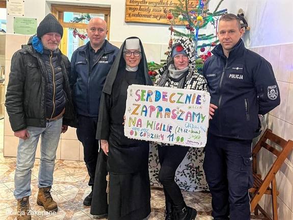 W korytarzu na tle choinki dwóch policjantów, zakonnica i para po cywilnemu z tabliczką zapraszamy na Wigilię