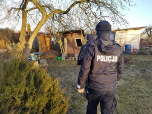 Widziana z tyłu dwójka policjantów na tle rozpadających się zabudowań gospodarczych