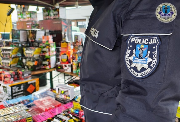 Zbliżenie torsu policjanta w kurtce służbowej na tle stoiska ze sztucznymi ogniami