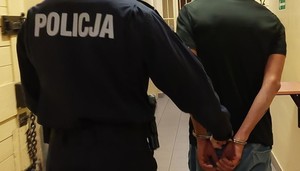 widziani z tyłu w korytarzu pomieszczeń dla zatrzymanych policjant przytrzymujący mężczyznę ze skutymi z tyłu rękami