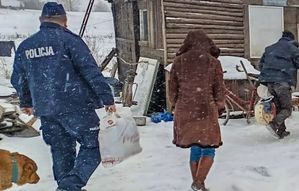 W scenerii zimowej widziani z tyłu dwaj policjanci z torbami oraz kobieta w kożuchu idący do małego budynku drewnianego