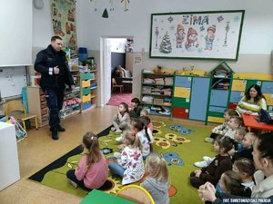 Policjant prowadzący pogadankę dla siedzących na wykładzinie sali przedszkolaków.