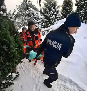 W scenerii zimowej policjant oraz ratownicy medyczni niosący na noszach mężczyznę